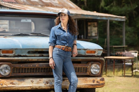 A white woman in denim shirt, jeans and a cap and long frizzy hair leans against a vintage pickup truck