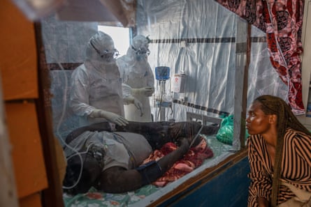 Two health workers tend to a man lying on a bed as a woman looks on through a transparent protective sheet