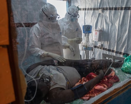 Two healthcare workers in full protective suits stand beside a patient lying on a hospital bed.