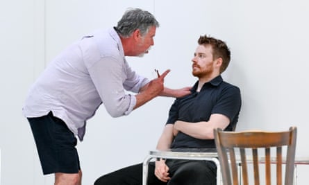 Jack Lowden is seated by a table. Sean Gilder leans over him, pointing at Lowden’s face and talking