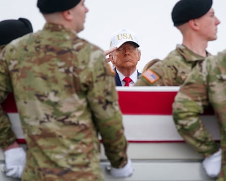 Trump, wearing a white cap, salutes, a soldiers carry a coffin draped with the US flag