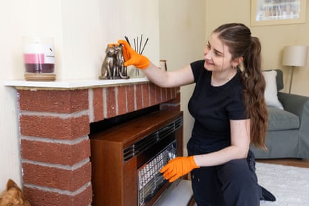 A woman wearing a black T-shirt and black trousers and orange gloves, smiling as she places two bronze cats on a fireplace.