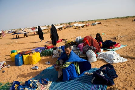 A man wrapped in a blanket sits on a plastic mat, on sand, with other people huddled nearby.