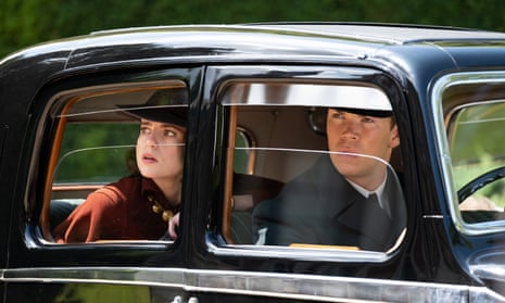 Lucy Boynton and Will Poulter in Why Didn’t They Ask Evans? in a 1930s car, wearing hats.