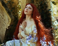 A woman with long red hair and a 19th-century dress sits next to a gravestone and flwers holding her hand and looking wistful