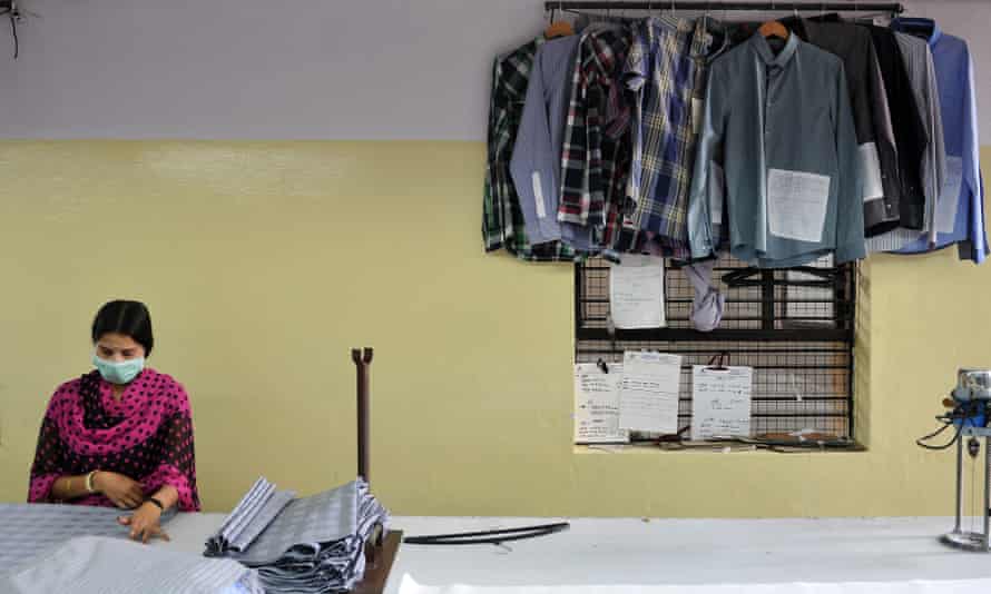 An Indian garment worker sits at a table sorting through clothing that is to be sewn together with shirts hanging up behind her