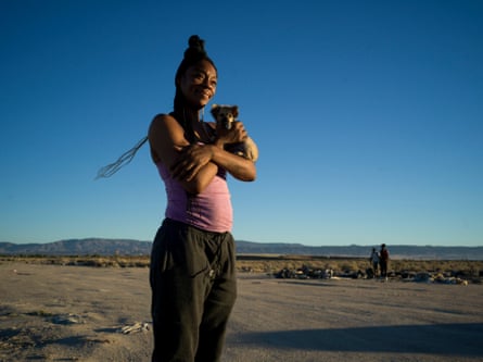 A woman with braids wearing a pink top holds her dog in the desert