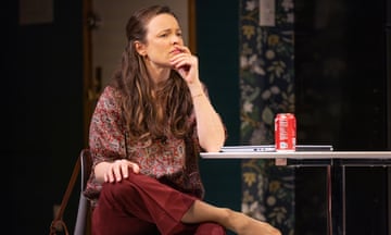 A white woman with long brown hair, dressed in a red paisley long-sleeved blouse and maroon pants, sits at a kitchen table in front of a can of Coke and a laptop, with her legs crossed and her hand on her knee, her chin resting in her other hand.