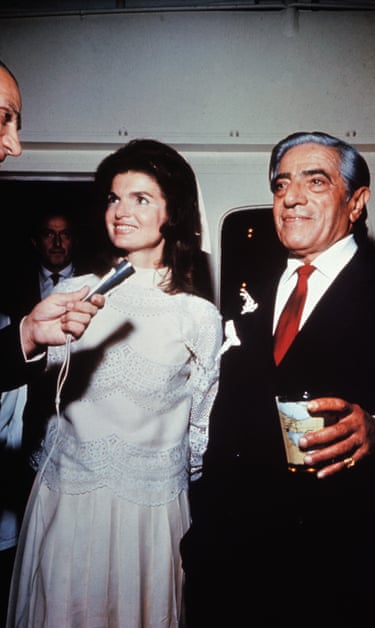 Jacqueline Kennedy in a white dress smiling with Aristotle Onassis as a reporter holds up a microphone