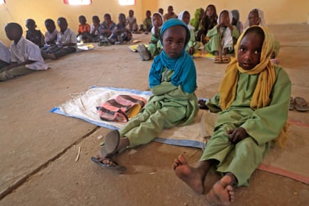 Two small girls look at the camera in a classroom full of children sitting on the floor
