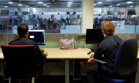 Two Border Force officers sit at computer screens behind windows looking out over people queuing at immigration control