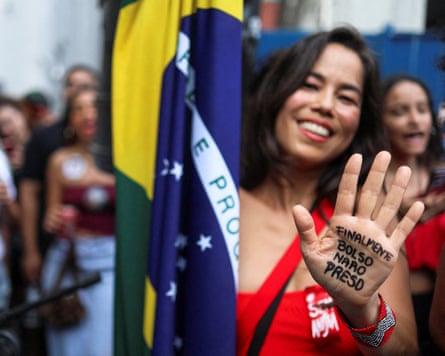A smiling woman with a flag holds up her hand, which has the words ‘Bolsonaro finally arrested’ written on it in Portuguese