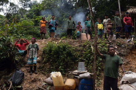 A group of men, women and children stand in a clearing in the jungle. Smoke rises from an unseen camp fire behind them