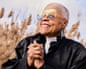 A woman with cropped white/grey hair, wearing large, orange transparent sunglasses and black and white leaf-shaped earrings, a white high-collared zip-up top and a shiny black coat, clasping her hands together and grinning off camera, standing against a background of a field of wheat