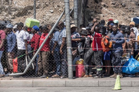 A queue of people with bags behind a wire fence