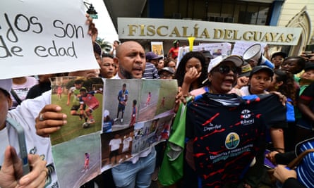 Luis Arroyo holds up photos of his sons playing football in a crowd in front of the prosecutor’s office; a football shirt is also held up and people behind are shouting and crying