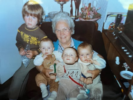 An elderly women sits on armchair holding baby triplets in her arms, with a young boy sitting next to them on the arm of the chair