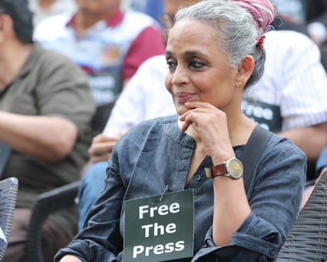 Arundhati Roy sits at a protest with a sign saying 'Free the press' attached to her shirt