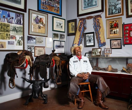 Black man in cowboy hat, white cowboy shirt, red scarf at his neck, leather chaps over jeans, and cowboy boots, sits back in a wooden chair, smiling, beside two saddles on display and a corner of a room covered in framed photographs.