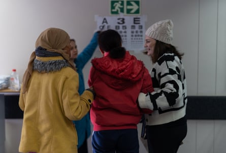 A girl is supported by two women as she stands in front of an eye chart on the wall