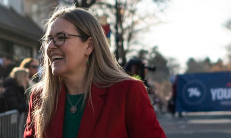 White woman with long blond hair, glasses, and red blazer laughs in afternoon sunlight on the side of a street where there is a parade behind her.
