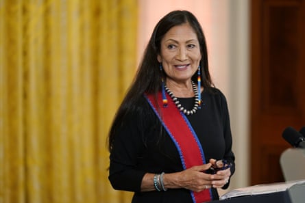 A woman in long beaded earrings, a heavy silver necklace, and silver and turquoise bracelets speaks at a low lectern.