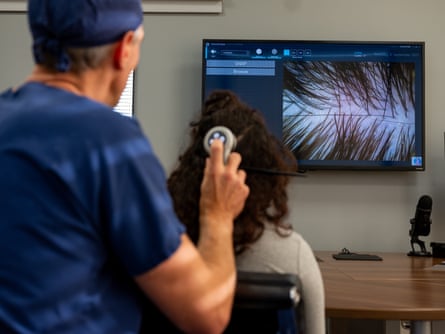 A man in a blue medical top and hat, holding a machine up to the back of the head of a woman looking at her own scalp on a screen