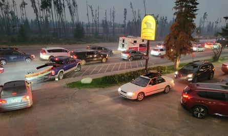 A queue of cars with headlights on leaving Jasper national park in Canada, with black trees silhouetted against a hazy sky as darkness falls in the background