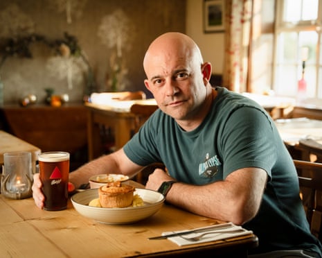 Dan Smith sits at a pub table. He has a pint of ale in his right hand and a shallow bowl with a pie on top of mashed potatoes sitting in front of him on the table.