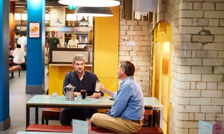 Two men, both wearing blue shirts, sitting at a restaurant table talking