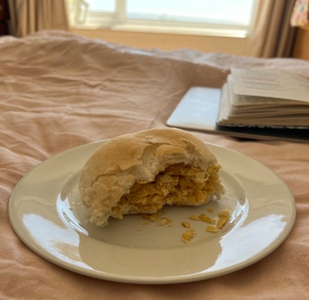 A white roll stuffed with crisps, on a plate, on the bed, with a book open for reading