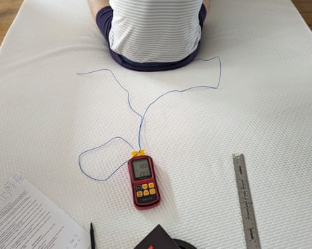 A person is at the top of the frame sitting in shorts in the centre of the mattress there is a gadget, pen, info sheel and metal rule displayed on the mattress also.