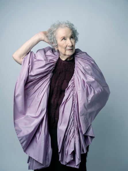 Margaret Atwood wearing a deep-purple fringe-knit outfit with a lilac cape-like shawl draped around her, standing with her left arm on her hip beneath the shawl and her right arm bent at the elbow with her hand on the back of her head