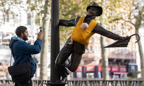 A man photographs a statue of Gene Kelly swinging from a lamppost with a model baby in a sling wrapped around