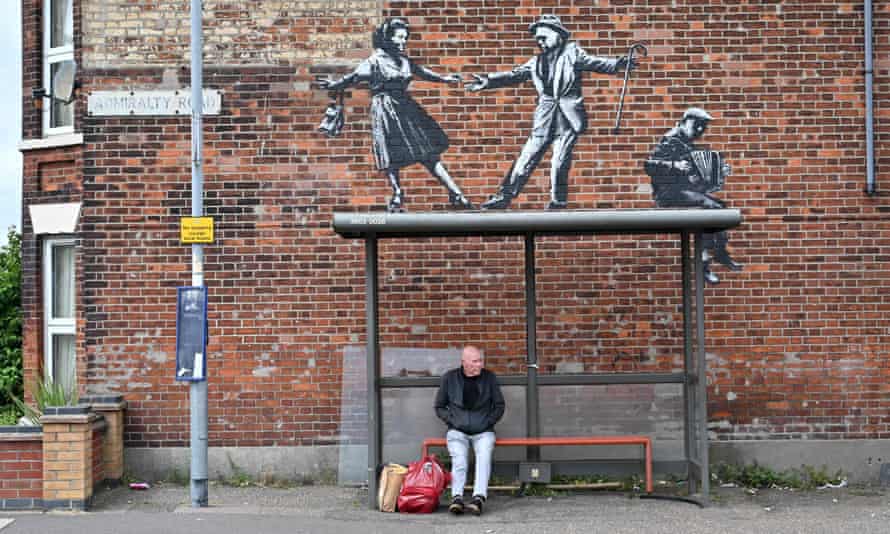 A man waits at a bus stop below a graffiti artwork of a couple dancing to an accordion player, by street artist Banksy, on a wall in Great Yarmouth
