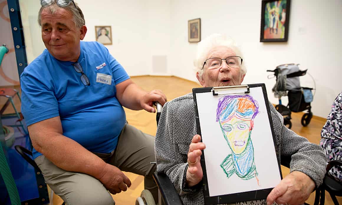 Harry Bruinenberg, in a blue shirt, sits next to his white-haired mother, Bwieukje Bruinenberg-Haisma, who is in a wheelchair and holding a colorful drawing of Alexej von Jawlensky’s Head of a Woman. Bwieukje is mouthing the word “mooi,” which means beautiful in Dutch.
