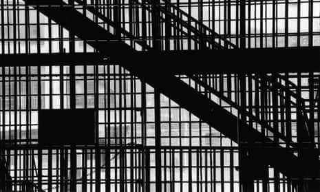 A close-up of vertical prison bars with a diagonal staircase with railing silhouetted against large windows in an abandoned structure.