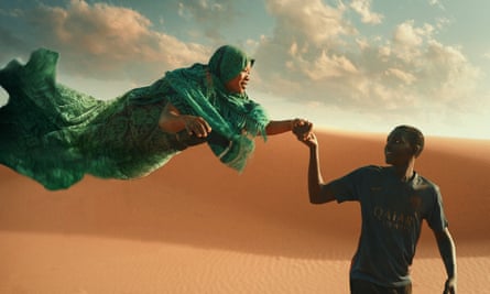 A young man in a Paris St-Germain shirt leading a floating woman in a rippling green robe across a desert