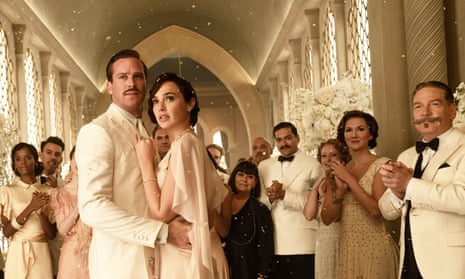 Actors Armie Hammer and Gal Gadot, dressed in a a 1930s white suit and white dress respectively, stand in the foreground in a still from Death on the Nile. The movie's other cast members stand in the background, also dressed primarily in light colors.