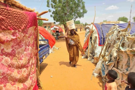 A woman walks carrying a plastic canister on her head between a line of makeshift tents and shelters that have been made from sacking, tarpaulins and blankets. She walks on yellow sand.