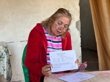 An older woman with glasses sits at a table holding a typed bit of paper to the camera