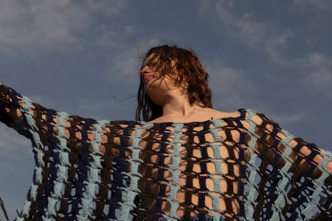 A model luxuriating in a blue and black crocheted jumper with their arms spread wide, against a blue-grey sky.