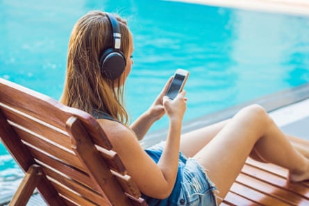 Woman relaxing on a sunbed next to a pool, with headphones on and a phone in her hands