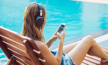 Woman relaxing near swimming pool listening with earbuds to streaming music and on a phone.
