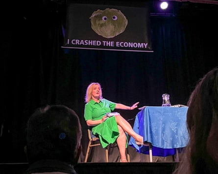 Liz Truss wearing a bright-green dress sitting at a table during a talk on a stage with a banner above her showing a googly-eyed lettuce with 'I crashed the economy' written underneath