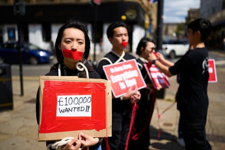 A line of young Chinese protesters on a street with red tape over their mouths. Chloe holds a sign saying “£100,000 – wanted”