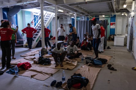 People stand and sit on towels below desk on a ship.