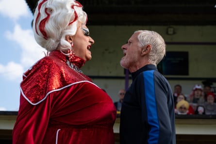 A man dressed up in drag with a silky and velvety red dress, a silver wig with red streaks. They are shouting at a man with grey hair and a beard, wearing a tracksuit.