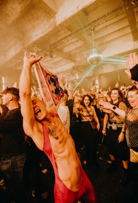 A bald man wearing a topless red unitard throws his arms in the air on a packed dancefloor