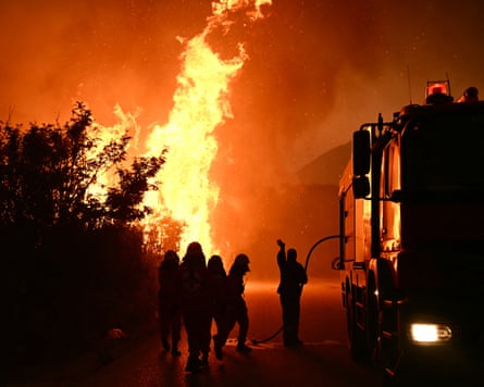 A night shot of a fire engine and firefighters in silhouette with flames leaping into the sky behind them.